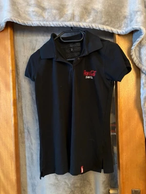 Svart pikétopp Coca-Cola Zero - Svart pikétopp med korta ärmar från Coca-Cola Zero. Toppen har klassisk krage, knappar framtill och broderad logga i rött och vitt på bröstet. Perfekt för dig som gillar unik merch och vill sticka ut lite extra.
