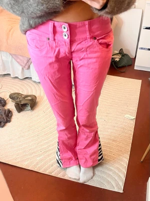 Rosa bootcut jeans med zebra-detalj - Unika rosa bootcut byxor med två knappar i midjan och coola zebramönstrade kilar längst ner på benen. Byxorna har normal passform och är perfekta för dig som vill sticka ut. Materialet känns som bomull och de har klassiska fickor fram.