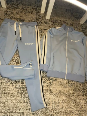  Palm Angels Tracksuit - Säljer ett ljusblått tracksuit-set från Palm Angels med vita och svarta ränder längs ärmar och ben. Setet består av en zip-jacka med hög krage och två fickor samt matchande byxor med dragsko i midjan och dragkedja vid benslut. Perfekt för chill dagar.