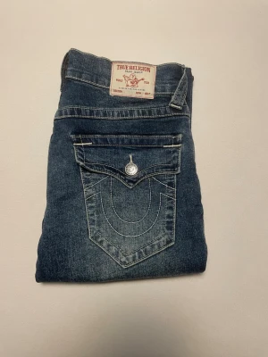 Blå jeans från True Religion W26 L32 - Snygga blå jeans från True Religion med klassiska kontrastsömmar och stora fickor med lock och knapp bak. Jeansen har en rak passform och är tillverkade i slitstarkt denimtyg. Perfekta för dig som vill ha en cool och tidlös look.