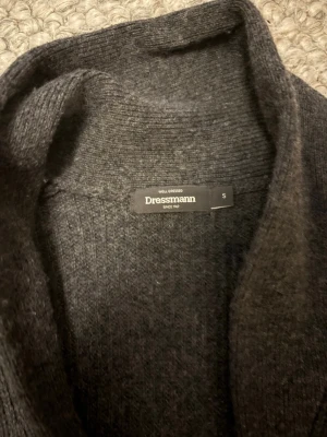 Mörkgrå kofta från Dressmann - Mysig mörkgrå kofta från Dressmann med ribbstickad struktur, knäppning framtill och två praktiska fickor. Perfekt för lager på lager och enkel att matcha med olika outfits. Klassisk och stilren design med mjukt material.