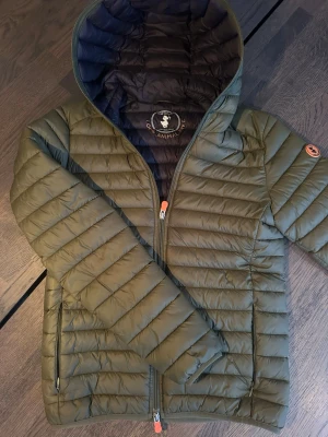 Save The Duck jacka - Snygg olivgrön dunjacka från Save The Duck med quiltad design och huva. Jackan är lättviktig, har dragkedja framtill och orange detaljer på zipparna. Märket är känt för sina djurfria material och jackan är perfekt för kyliga dagar.                                              Nypris: 1999kr