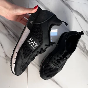 Svarta sneakers  Emporio Armani EA7 - Snygga svarta sneakers från Emporio Armani EA7 med meshdetaljer och logga på sidan. Skorna har snörning, platt sula och sportig design. Inga defekter, sidan på sulan är bara smutsig. Min bror växte är de snabbt så de blev knappast använda. Köptes för 2200kr. Kom med prisförslag vid snabb affär🥰