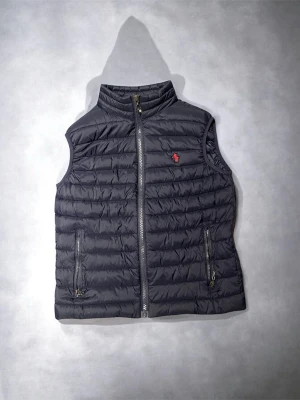 Mörkblå väst från Polo Ralph Lauren - Snygg mörkblå väst från Polo Ralph Lauren med ståkrage och dragkedja framtill. Västen har två fickor med dragkedja och den klassiska röda loggan broderad på bröstet. Perfekt för lager på lager och ger en sportig vibe. Västen är i strl m men passar bättre s. (vet ej om den är original)