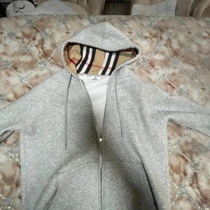 Grå hoodie från Burberry med dragkedja - Snygg grå hoodie från Burberry med klassiskt rutigt foder i huvan. Tröjan har dragkedja framtill, snörning i huvan och broderad Burberry-logga på bröstet. Perfekt för dig som gillar stilrena och exklusiva plagg storlek m men är liten i storleken dragkedjan har gått sönder men det går att stänga tröja. Pris kan absolut diskuteras