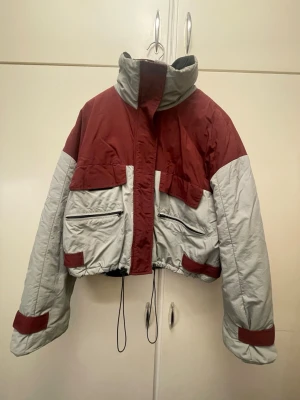 Röd och grå pufferjacka med hög krage - Snygg pufferjacka från Gina i rött och grått med hög krage och dragsko längs ned! Jackan har flera fickor och kardborreband vid ärmslut. Perfekt för våren och hösten och ger en cool streetstyle-look!