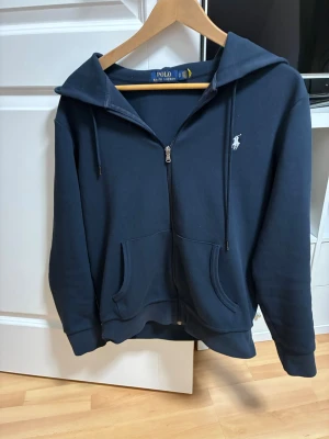 Mörkblå hoodie från Polo Ralph Lauren - Mörkblå hoodie med dragkedja från Polo Ralph Lauren. Klassisk design med vit broderad logga på bröstet, huva med dragsko och två fickor framtill. Perfekt för en avslappnad och stilren look.