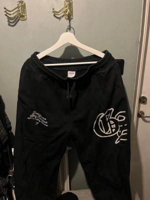 Svarta mjukisbyxor med tryck från Gymshark  - Svarta mjukisbyxor från Gymshark med resår och snörning i midjan. Stora vita tryck på ena benet och mindre texttryck på andra sidan framtill. Perfekta för en chill och avslappnad stil.