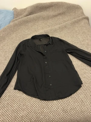 Svart skjorta med bröstficka - Stilren svart skjorta med klassisk krage, knäppning framtill och en bröstficka. Skjortan har långa ärmar med breda manschetter och är tillverkad i ett mjukt, tunt material som ger en avslappnad passform. Perfekt för en clean och enkel look. Oklar storlek och märke då lappen är gammal, men den sitter som en S. 