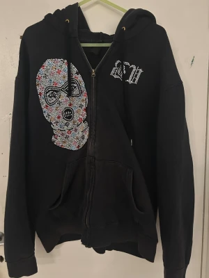 Svart hoodie med strassmask och dragkedja - Svart hoodie med dragkedja och stor ficka framtill. På vänster sida finns en stor maskdesign i färgglada strasstenar och på bröstet står det 'MSW' i strass. Hoodien har huva med snören och är gjord i mjukt bomullsmaterial. Perfekt för dig som gillar streetwear och unika detaljer.