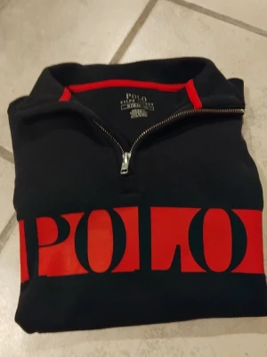 Svart tröja från Polo Ralph Lauren - Svart tröja från Polo Ralph Lauren med stor röd POLO-logga över bröstet. Tröjan har även en liten polo logga broderad på ena armen. Tröjan är väldigt sparsamt använd. Tröjan är en klassisk half-zip och är perfekt för dig som gillar att ha en avslappnad samt snygg stil.