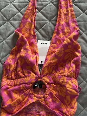 Blommig topp - Säljer en färgstark topp från Bik Bok med rosa och orange blommönster. Toppen har djup v-ringning, ringdetalj framtill och breda axelband. Perfekt för sommarens alla tillfällen