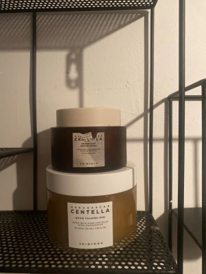 SKIN1004 Centella ansiktsprodukter - Två ansiktsprodukter från SKIN1004: en brun burk med beige lock och en större transparent burk med vitt lock. Båda tillhör Centella-serien, varav den ena är Quick Calming Pad. Perfekt för dig som gillar koreansk hudvård.