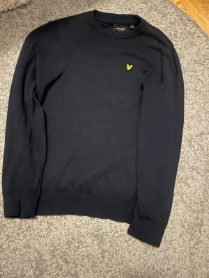 Svart stickad tröja från Lyle & Scott - Klassisk svart stickad tröja från Lyle & Scott med rund hals och gul broderad logga på bröstet. Tröjan har lång ärm och ribbade muddar vid ärmslut och nederkant. Perfekt för en stilren look.