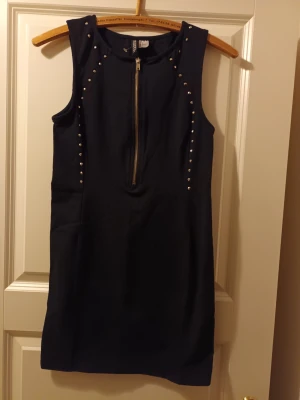 Svart ärmlös klänning med nitar - Snygg svart kortklänning med silverfärgade nitar längs axlar och byst. Klänningen är ärmlös och har en dragkedja framtill som detalj. Perfekt för dig som gillar en edgy och stilren look.