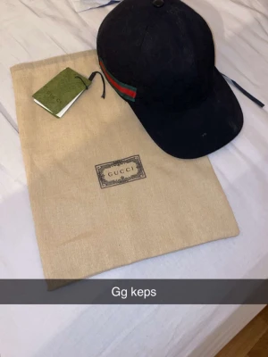 Svart keps från Gucci med GG-mönster - Säljer en svart keps från Gucci med det klassiska GG-monogrammet och gröna/röda ränder på sidan. Kepsen har böjd skärm och justerbar rem bak. Kommer med originalpåse och box. Perfekt för dig som vill ha en stilren och lyxig accessoar.
