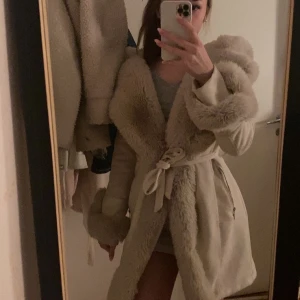 Beige kappa med fuskpäls från Garoff - Elegant beige kappa från Garoff med stora detaljer i fuskpäls på krage, ärmslut och längs öppningen. Kappan har knytband i midjan för en markerad siluett och är långärmad. Perfekt för kalla dagar när du vill vara både varm och stylish.