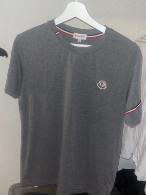 Moncler t-shirt - Snygg grå t-shirt från Moncler med klassiskz rund hals och korta ärmar. Diskret Moncler-logga på bröstet och cool detalj med röd, vit och blå rand på ärmslutet. Perfekt till jeans eller shorts för en clean look. Moncler märket på bröstet sitter lite löst men det går att fixa med några nya stygn så inga problem! Tveka inte på att ställa en fråga eller två!🤝🥰