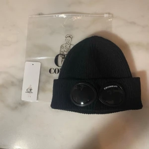C.P CompanyMössa - Svart stickad mössa med goggles framtill – clean, modern och lätt att matcha med allt. Perfekt för både vardag och outfits. Skön kvalitet med bra stretch, sitter stabilt och bekvämt. Kommer med originalpåse och tag. Onesizestorlek.  Mycket bra skick.  📦 Fraktar direkt efter köpt 💬 Pris kan diskuteras 🔥 Rabatt vid köp av båda