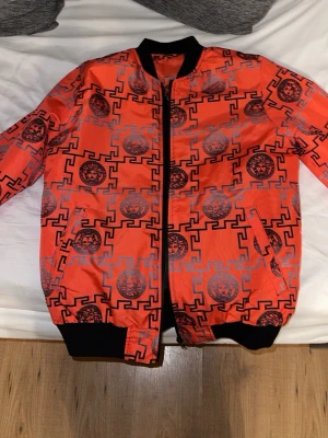 Orange bomberjacka med grafiskt mönster - Snygg orange bomberjacka med svart och grått grafiskt mönster och Medusa-liknande tryck. Jackan har svarta muddar vid ärmslut, krage och nederkant samt dragkedja framtill och två fickor. Perfekt för dig som vill sticka ut med en unik streetwear-look.