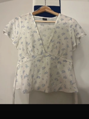 Blommig omlottopp från Gina Tricot - Söt vit omlottopp från Gina Tricot med ljusblått blommönster. Toppen har korta ärmar, v-ringning och knytband i midjan. Perfekt för en fräsch och somrig look.