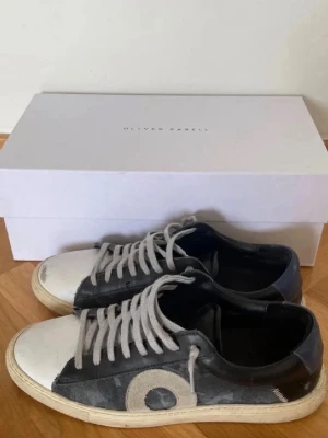 Svarta sneakers från Oliver Cabell - Snygga svarta sneakers från Oliver Cabell med vit tå och sula. Skorna har blå mockadetaljer och ett stort O på sidan. De har vita skosnören och en klassisk låg profil. Materialmix av läder och mocka ger en stilren och modern look. Skorna passar upp till 41