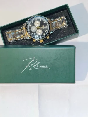 Pelvini klocka Dominate Obsidian - Stilren klocka från Pelvini med metallarmband i silver och gulddetaljer. Urtavlan är svart med tre mindre vita subdials och guldfärgade knappar. Klockan har en klassisk rund form och levereras i original grön box.                  Nypris 1500!                                              Skicka ett bud så löser vi ett bra pris. Hämtas upp i Nacka samt kan mötas i nästan hela Stockholm eller frakta!