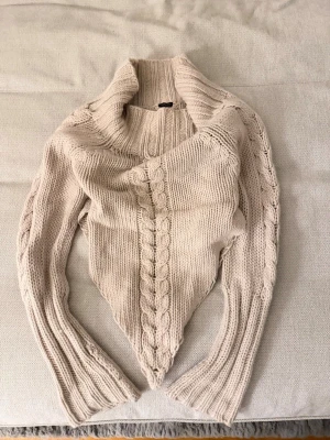 Beige stickad tröja med kabelmönster - Säljer en beige stickad tröja med bred polokrage och snyggt kabel sopmönster framtill och längs ärmarna. Tröjan är gjord i en mjuk blandning av ull och akryl, vilket gör den både varm och skön. Perfekt för kyliga dagar och enkel att styla till jeans eller kjol.