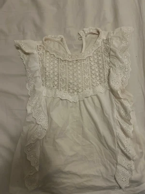 Vit spetsblus med volangdetaljer - Söt vit blus med broderade spetsdetaljer och volanger längs ärmöppningarna. Blusen har ett vackert hålmönster framtill och knytning i nacken. Perfekt för dig som gillar romantisk och bohemisk stil. Tillverkad i bomull eller liknande material.