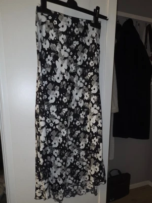 Svart långkjol med blommigt mönster - Snygg svart långkjol med vita och grå blommor. Kjolen har en rak och flowy passform som ger en avslappnad men ändå stilren look. Perfekt för dig som gillar blommiga prints och vill ha något som sticker ut lite extra.