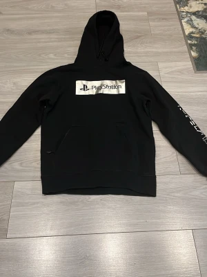 Svart PlayStation hoodie med tryck - Snygg svart hoodie från PlayStation med stort silverfärgat PlayStation-tryck på bröstet och text på ena ärmen. Klassisk känguruficka framtill och justerbar huva med snören. Perfekt för dig som gillar gaming och vill ha en stilren look.