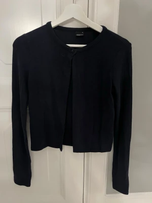 Svart kofta med rund hals - En stilren svart kofta från Gina med rund halsringning och lång ärm. Koftan har en enkel knäppning upptill och är tillverkad i ett mjukt, tunt material som passar perfekt till lager-på-lager-stil.