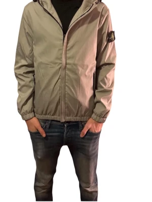 Beige vindjacka från Stone Island - Säljer en beige vindjacka från Stone Island med huva och dragkedja framtill. Jackan har svarta detaljer, elastiska muddar och klassisk Stone Island-patch på vänster ärm. Perfekt för dig som vill ha en stilren och sportig look.