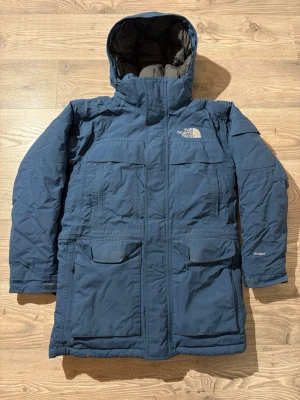 The North Face - Blå parkasjacka från The North Face med huva och flera stora fickor framtill. Jackan har lång ärm, diskret logga på bröstet och HyVent-tyg som står emot vind och väta. Perfekt för kalla dagar och outdoor-äventyr.