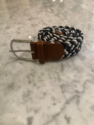 Flätat svartvittgrått bälte med läderdetalj - Snyggt flätat bälte i svart och vitt och grått med bruna läderdetaljer vid spännet. Spännet är i silverfärgad metall och bältet har en flexibel, elastisk känsla. Perfekt för att ge din outfit en cool touch.