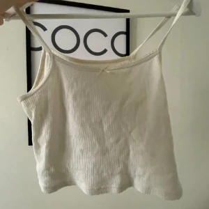 Gulligt linnetopp från Brandy Melville - Supersöt ribbad linnetopp från Brandy Melville i ljusvit färg. Toppen har smala axelband och en liten rosett framtill som detalj. Liten rosa aktig fläck som knappast syns, skriv för fler bilder!