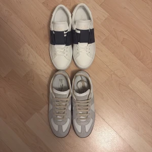 Valentino och Maison Margiela sneakers - Två par sneakers: Ett par vita Valentino Open med bred svart rem över vristen och klassisk logga, samt ett par Maison Margiela i vitt och beige med grå mockadetaljer och beige snörning. Båda paren har platt sula och rund tå för en clean och stilren look.
