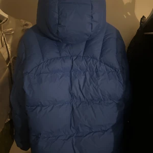 Blå dunjacka från Fjällräven - Säljer en blå dunjacka från Fjällräven med stor huva, gula snören och tryckknappar framtill. Jackan har två stora fickor med lock och Fjällrävens klassiska röd-gula logga på bröstet. Perfekt för kalla dagar och har en riktigt schysst puffer-look. Andvänd endast 1 vinter och är helt hel, inga märken eller hål på jackan, kemtvättas innan jag skickar iväg den. 