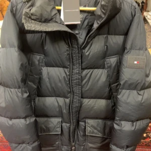 Svart pufferjacka Tommy Hilfiger S - Svart pufferjacka från Tommy Hilfiger i storlek S. Jackan har en hög krage, två stora fickor framtill och en snygg logotyp på ärmen. Materialet är polyester och dun, med quiltad design och dragkedja framtill. Perfekt för kalla dagar och riktigt stilren look.