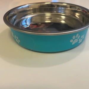 Praktisk och snygg kattmatskål! - Detta är en matskål för husdjur i rostfritt stål med blå utsida och vita tassavtryck. Skålen har en halkfri botten i svart gummi och är i gott skick. Perfekt för både mat och vatten till hund eller katt. Använts endast två gånger när vi har haft en kattpassning. 