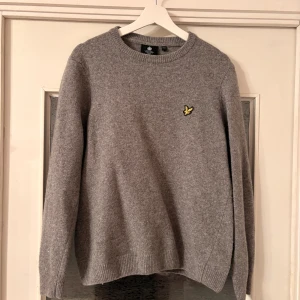 Grå stickad tröja från Lyle & Scott - Snygg grå stickad tröja från Lyle & Scott med rund hals och deras klassiska gula fågel-logga broderad på bröstet. Tröjan har ribbade muddar vid ärmslut och nederkant, och är långärmad. Perfekt för dig som gillar stilren och enkel design.