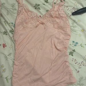 Ljusrosa spets linnetopp med rosett - Supersöt ljusrosa linnetopp med smala axelband och spets upptill. Toppen har en liten rosett i mitten och blommig spetsdetalj vid bysten. Materialet är mjukt och stretchigt, perfekt för en chill look. Sitter tight och har en v-ringad form.