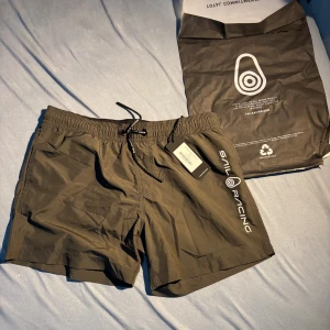 Mörkgrå Sail Racing shorts med snörning - Snygga mörkgrå shorts från Sail Racing, modell JR Bowman Volley. De har elastisk midja med snörning, logotryck längs benet och en bakficka. Materialet är lätt och snabbtorkande polyester, perfekt för strand eller pool. Stilren design med sportig vibe.