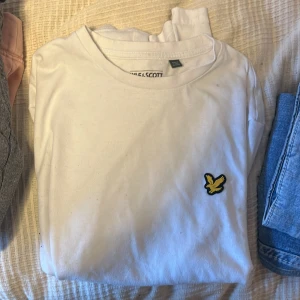 Vit t-shirt från Lyle & Scott - Vit t-shirt från Lyle & Scott med klassisk rund hals och korta ärmar. På bröstet sitter den ikoniska gula fågeln som broderad detalj. T-shirten är gjord i mjuk bomull och har en clean, stilren look som funkar till jeans eller shorts.