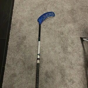 Unihoc Carbskin Slim 29 96cm - Unihoc Carbskin Slim 29 innebandyklubba med blått blad. Lätt och slitstark kolfiberkonstruktion, 29 flex. Greppet och bladet är i gott skick med minimalt slitage. Perfekt för både träning och matchspel.