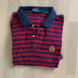 Randig pikétröja från Polo Ralph Lauren - Snygg pikétröja från Polo Ralph Lauren i storlek XXL. Tröjan har röda och marinblå ränder, klassisk krage och två knappar framtill. På bröstet finns ett broderat emblem i guld och rött. Perfekt för dig som gillar preppy stil och vill sticka ut.