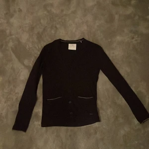 Svart kofta från EDC med knappar - Svart kofta från EDC med 5 knappar framtill och diskreta fickdetaljer. Långärmad och med v-ringad hals. Snygg och enkel design som passar till många olika stilar. Materialet känns mjukt och stretchigt, perfekt för lager-på-lager. 