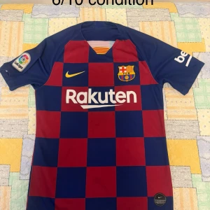 FC Barcelona matchtröja Nike rutig - FC Barcelona matchtröja från Nike med rutigt mönster i mörkblått och vinrött. Kortärmad modell med klubbmärke, La Liga-patch och sponsorer på bröst och ärm. Tillverkad i lätt och ventilerande material, perfekt för fotboll eller supporterliv.