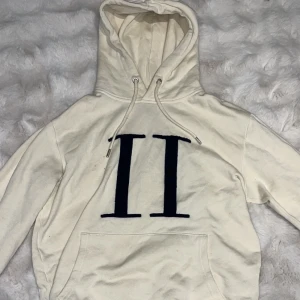 Les Deux hoodie  - Säljer en snygg vit hoodie från Les Deux med stort svart 'II'-tryck på bröstet. Tröjan har huva med snörning, känguruficka och är tillverkad i mjuk bomull. Perfekt för dig som gillar clean och stilren streetwear.