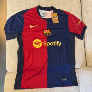 FC Barcelona matchtröja Nike röd/blå - Säljer en officiell FC Barcelona matchtröja från Nike med klassisk röd och blå halvdelad design. Tröjan har klubbmärke, Nike-logga och Spotify som sponsor framtill, samt UNHCR-tryck bak. Tillverkad i lätt och ventilerande polyester, perfekt för fotbollsälskare.